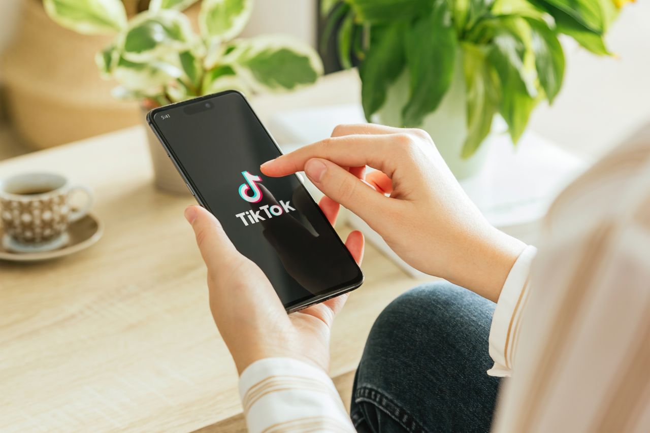 A person tapping the TikTok app on a smartphone while seated at a wooden table with a coffee cup and houseplants in the background.